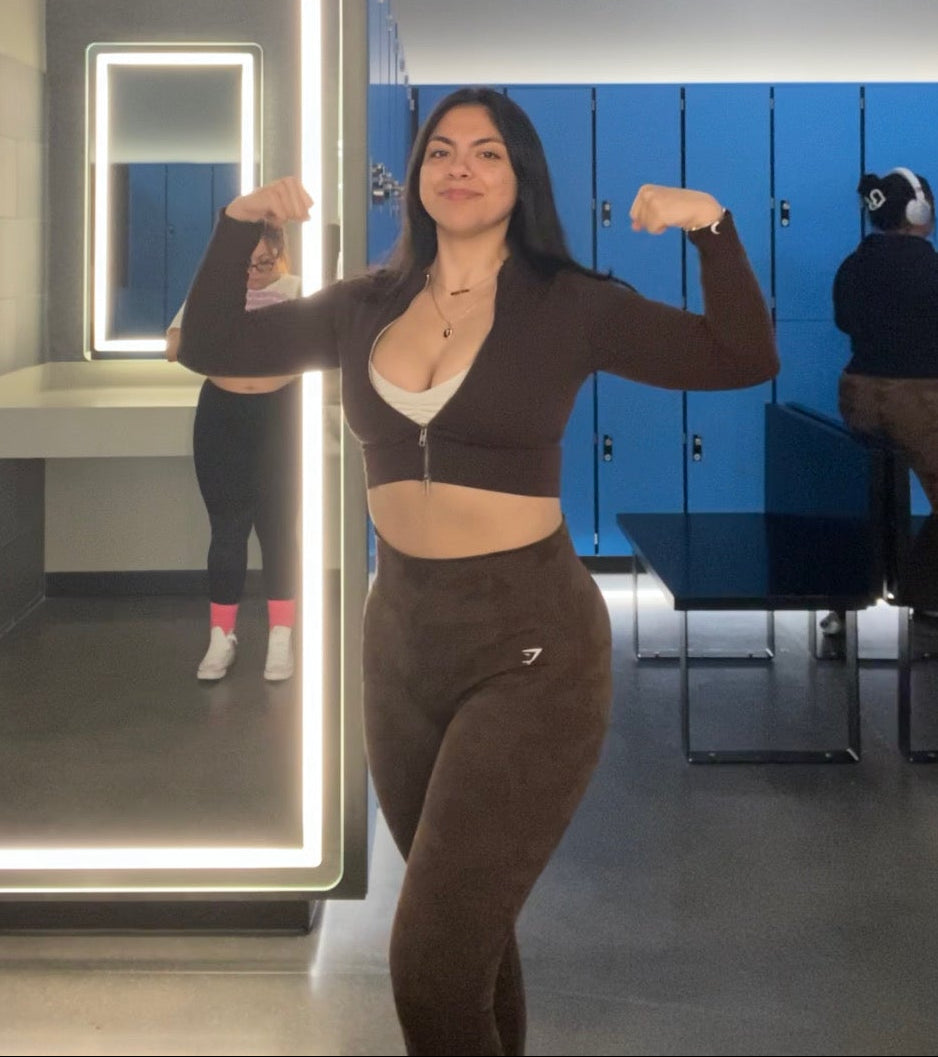 Woman flexing muscles in front of a mirror with a lit frame in a locker room.